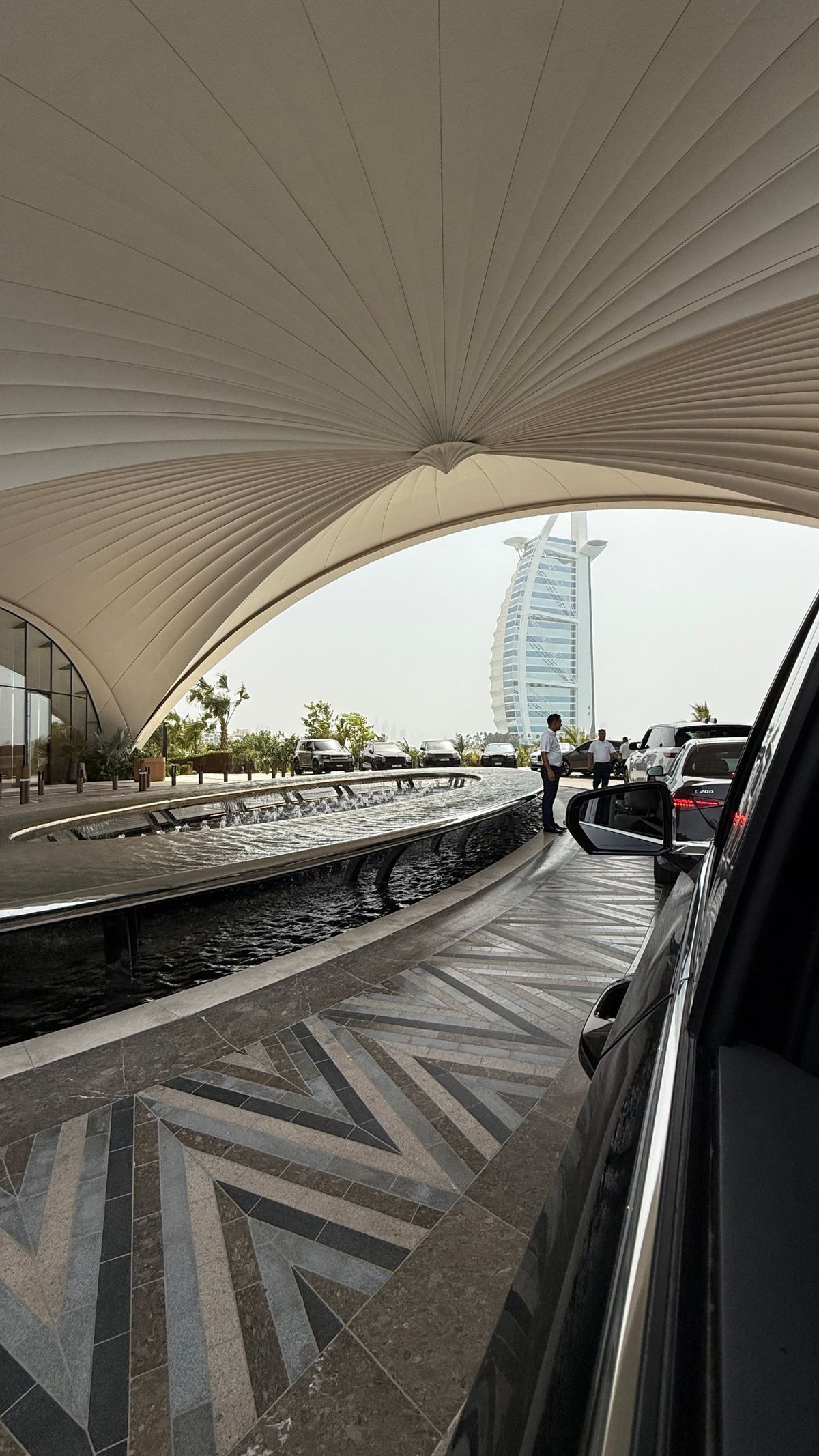 A hotel arrival court with a fountain, valets and a sail-shaped tower beyond