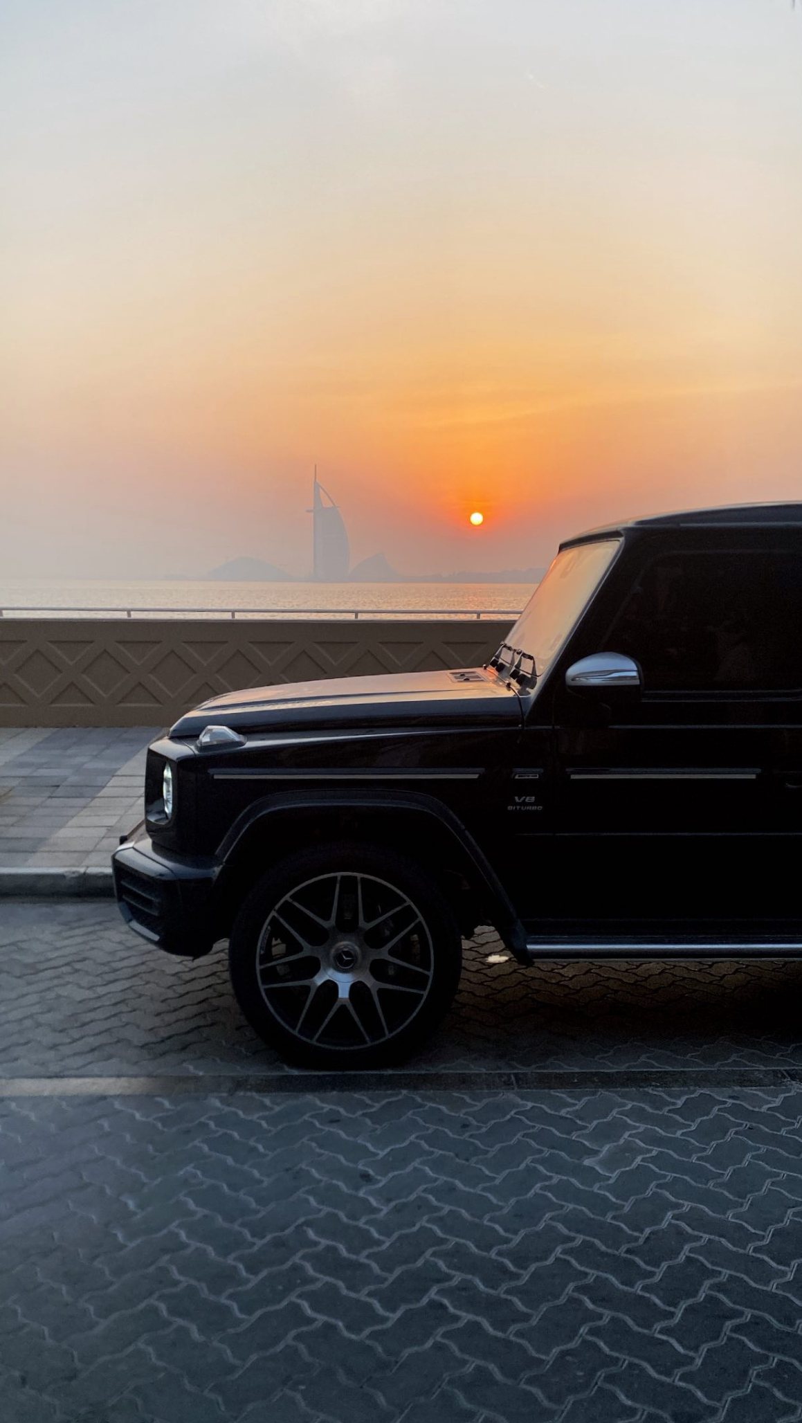 A black G-Wagon parked on a seafront promenade at sunset, a sail-shaped tower on the horizon